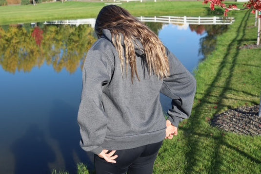 Person wearing a gray hoodie standing by a pond with trees reflecting on the water