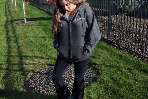 Woman in a gray hoodie standing by a pond with red leafless trees in the background.