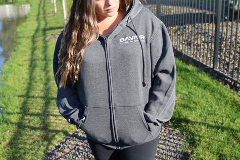 Person wearing a gray hoodie with a logo, standing outdoors by a body of water.