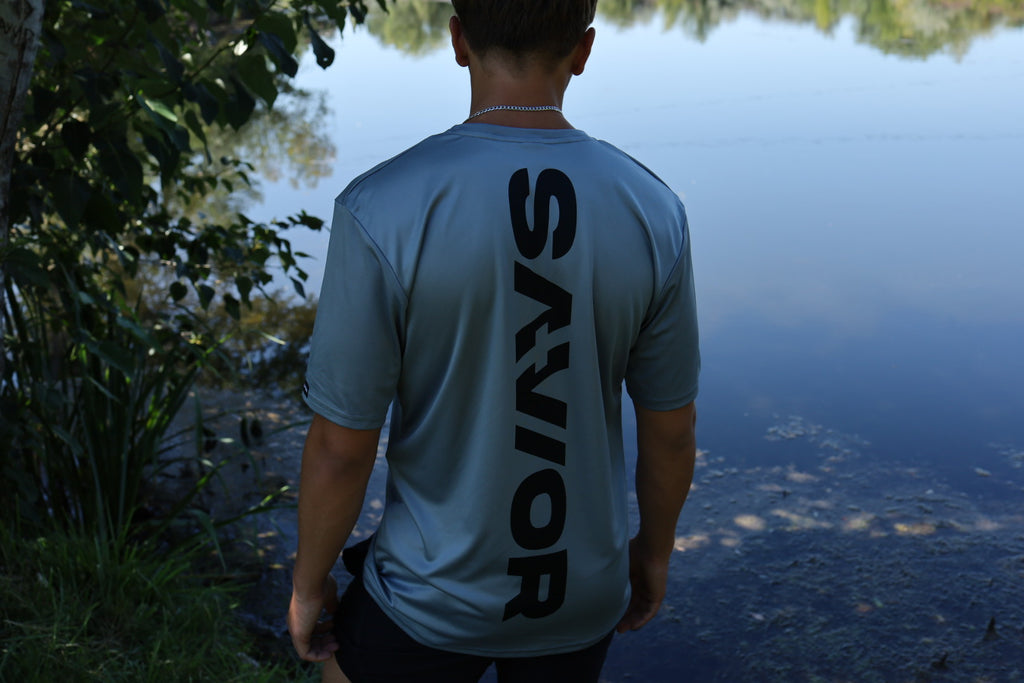 Person wearing a light blue t-shirt with 'SAVIOR' on the back, standing by a body of water.