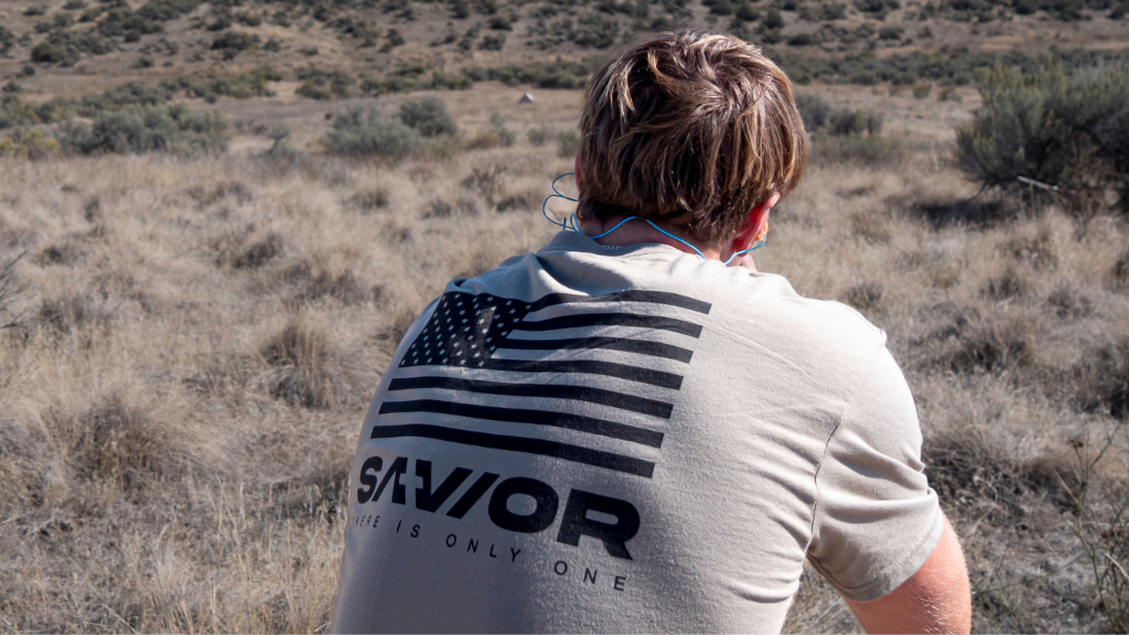Person wearing a shirt with 'SAVIOR' text and an American flag design in a desert landscape