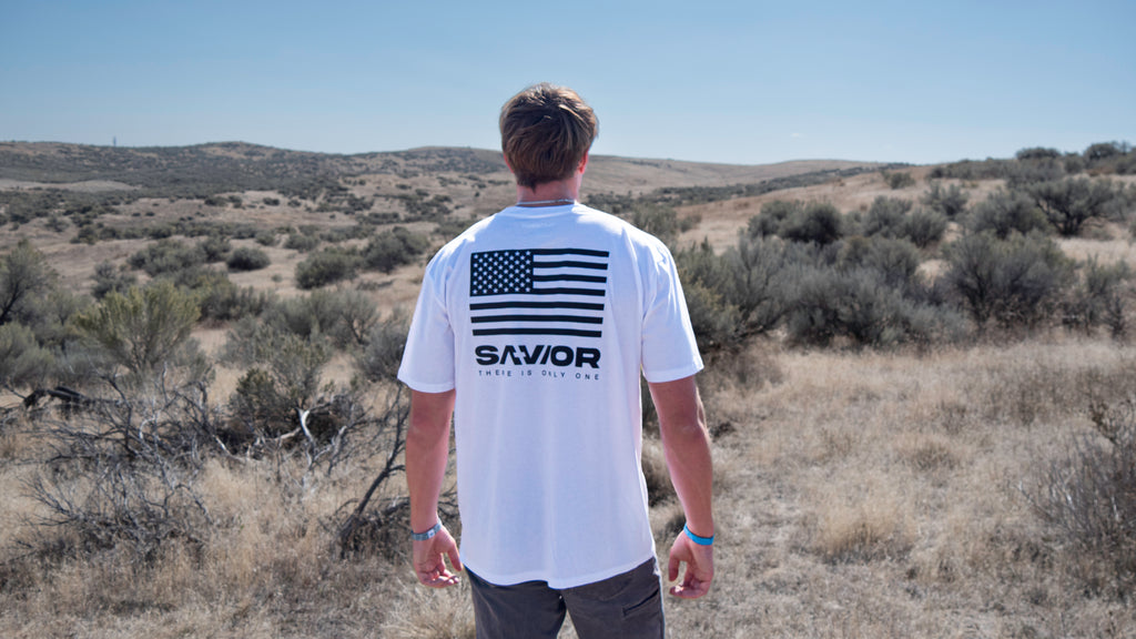 Person wearing a white t-shirt with an American flag design and 'SAVIOR' branding in a desert landscape.